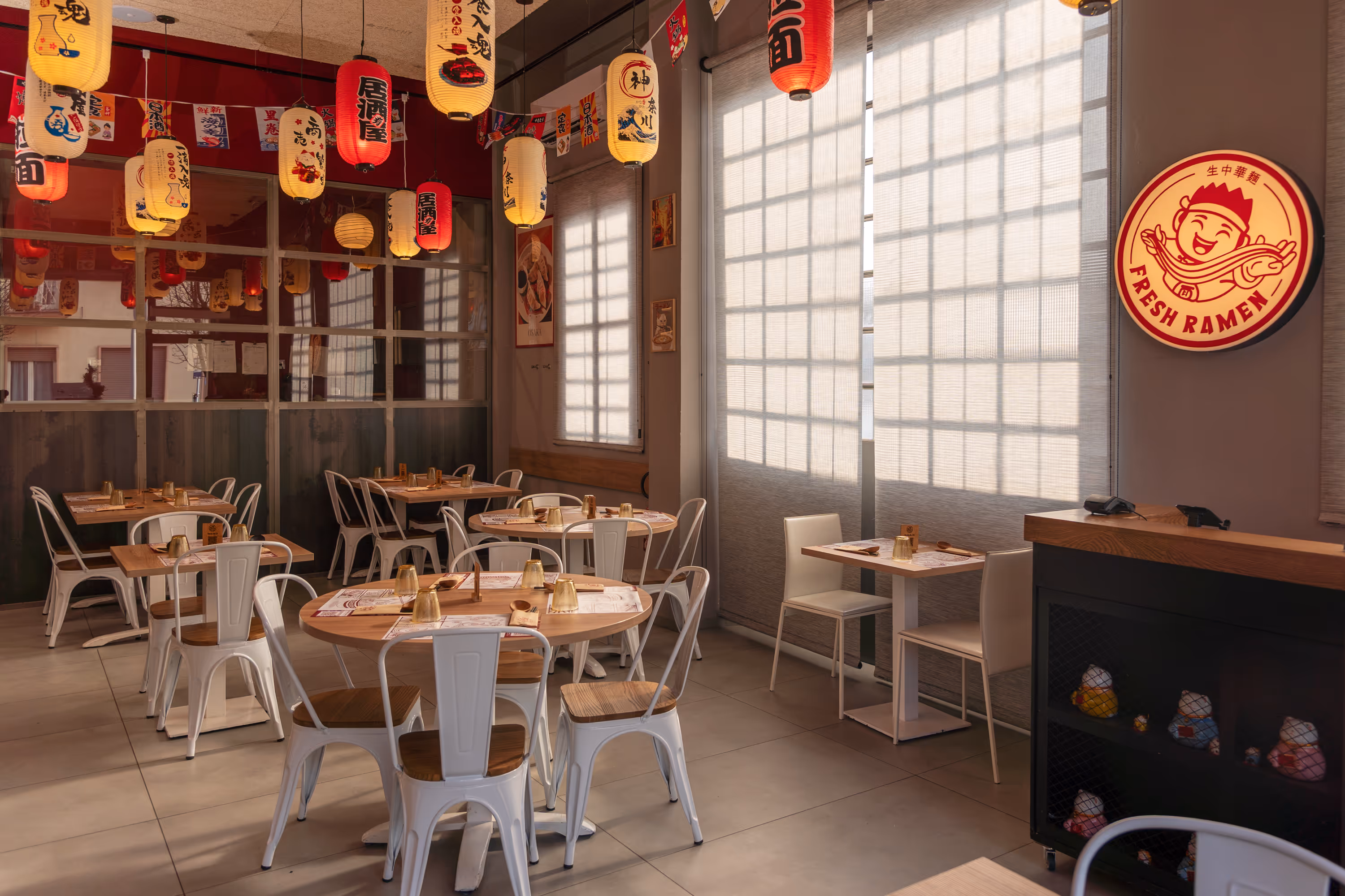Cozy ramen restaurant interior with wooden tables, white chairs, hanging Japanese lanterns, and a bright Fresh Ramen sign.