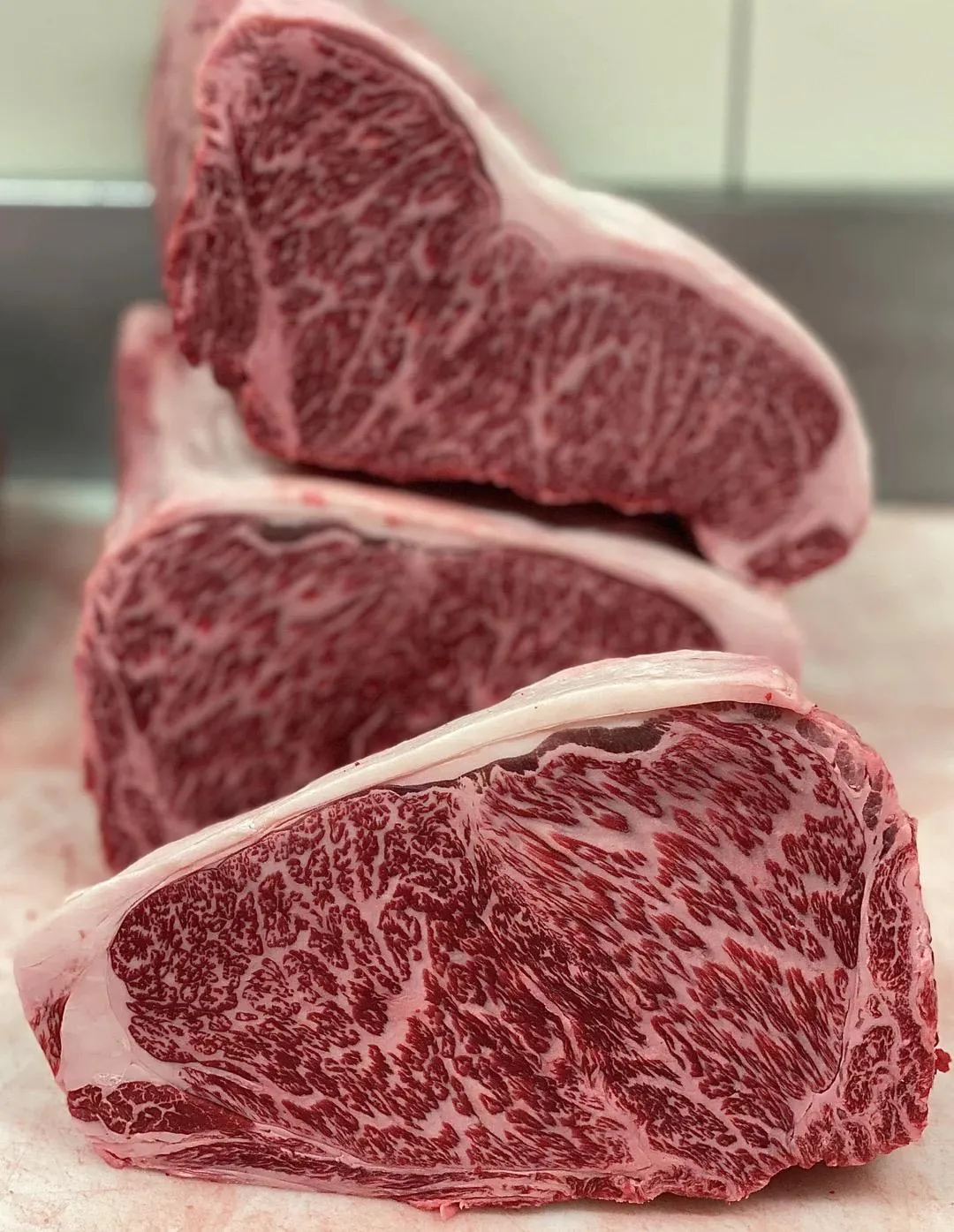 Three thick cuts of highly marbled raw beef steak stacked on a surface.