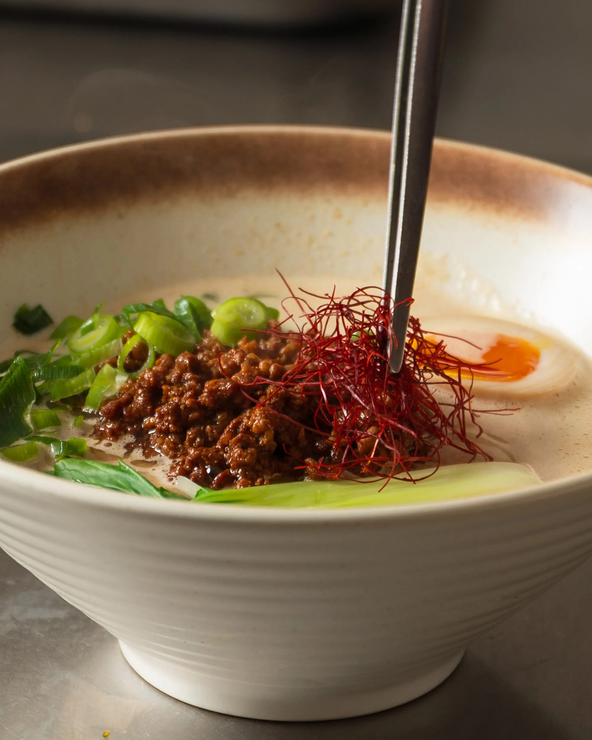 Bowl of ramen with ground meat, sliced green onions, soft-boiled egg, greens, and red chili threads being placed with chopsticks.