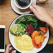 Plate with scrambled eggs, guacamole, greens, sliced sweet potatoes, and beets on a wooden table next to a coffee cup and smartphone.