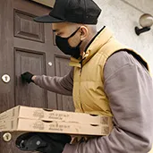 Delivery person wearing a black cap and face mask, holding two pizza boxes and knocking on a front door.