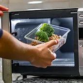 Person placing a plastic container with broccoli and minced meat into a microwave oven.