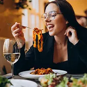 Woman with glasses smiling while holding a fork with spaghetti over a plate at a restaurant.