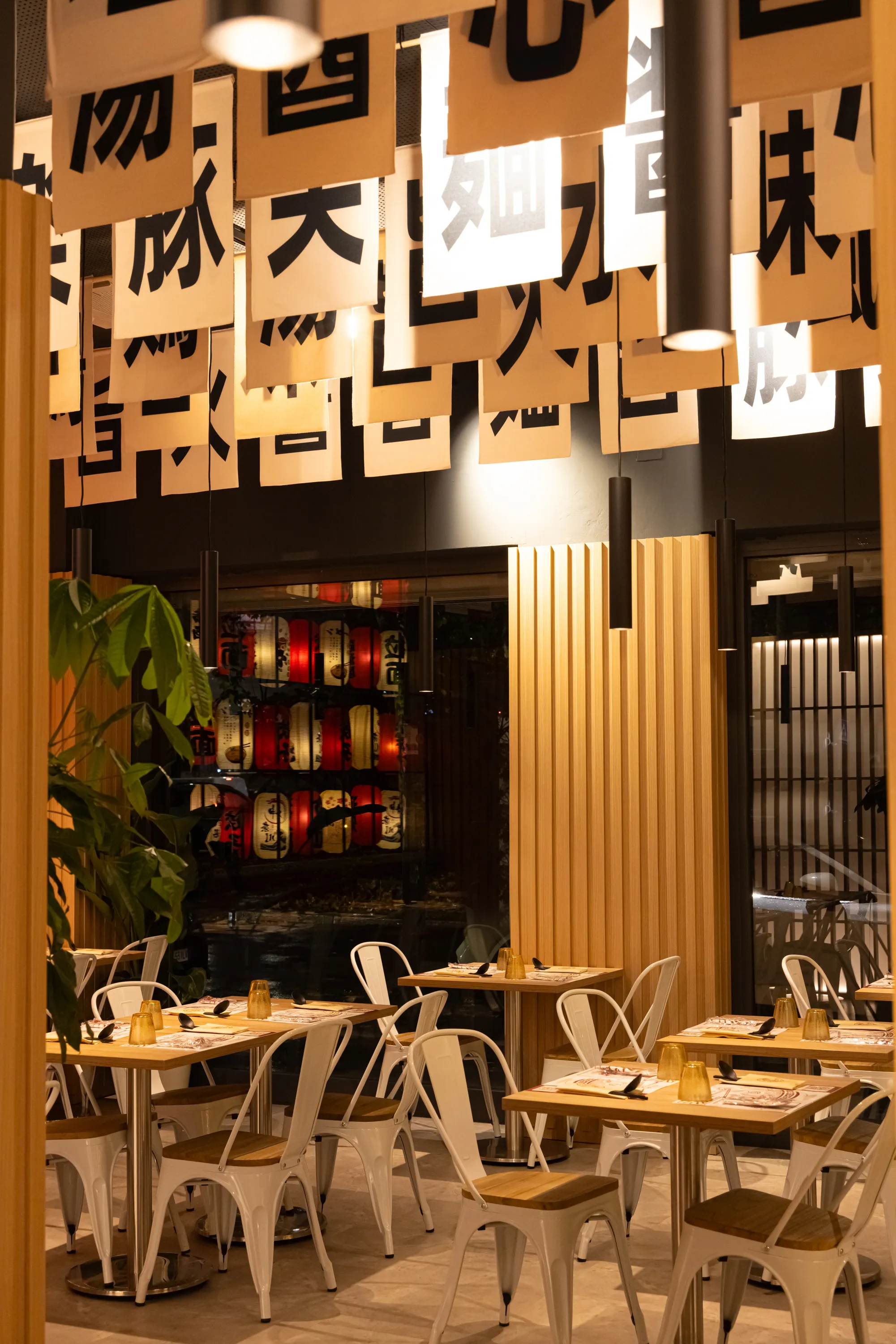 Interior of a modern restaurant with wooden tables, white chairs, hanging black pendant lights, and Japanese-style banners with black characters.
