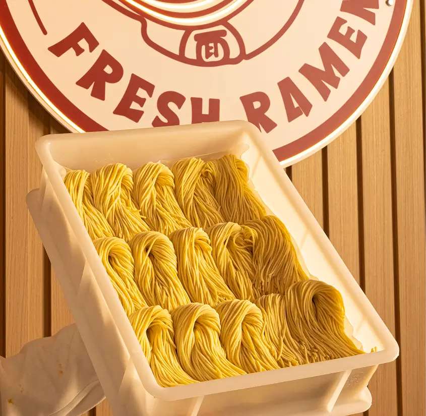 Tray holding neatly arranged bundles of fresh yellow ramen noodles in front of a wooden wall with a round sign reading 'Fresh Ramen.'