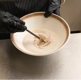 Hands wearing black gloves stirring a creamy mixture in a white bowl with a brown rim on a stainless steel surface.