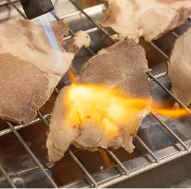 Thin slices of meat being seared with a flame on a metal grill.