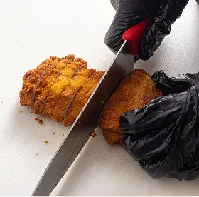 Person wearing black gloves slicing crispy fried chicken pieces on a white surface with a large knife.