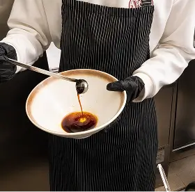 Person wearing black gloves and a striped apron pouring dark sauce into a white bowl.