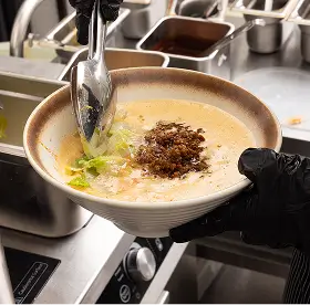 Person wearing a black glove holding a bowl of creamy soup topped with minced meat and garnished with chopped greens in a kitchen setting.