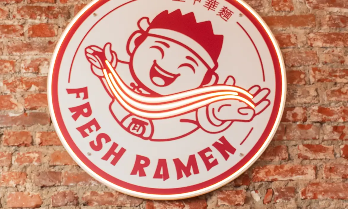 Round neon sign on brick wall featuring a smiling cartoon chef holding glowing ramen noodles with the text 'FRESH RAMEN.'
