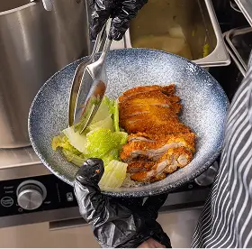 Crispy fried chicken served with lettuce in a blue ceramic bowl, held by a person wearing black gloves and a striped apron.