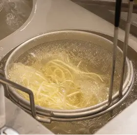 Pasta noodles boiling in a pot of water with a strainer inside the pot.