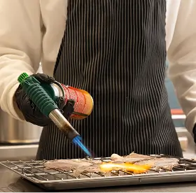 Person wearing black gloves and a striped apron using a small blowtorch to caramelize food on a wire rack.