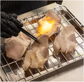 Person using tongs to sear thin slices of meat over an open flame on a wire grill.