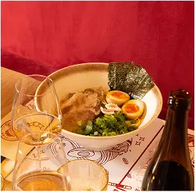 Bowl of ramen with sliced pork, soft-boiled eggs, green onions, and seaweed on a red surface beside wine glasses and a bottle.