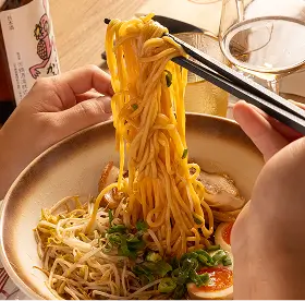 Hands holding chopsticks lifting noodles from a bowl of ramen with egg, bean sprouts, and green onions.