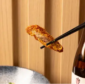 Chopsticks holding a piece of crispy fried food above a plate with a bottle nearby.