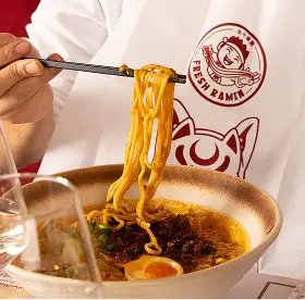 Hand lifting ramen noodles with chopsticks from a bowl containing broth, egg, and toppings on a table.