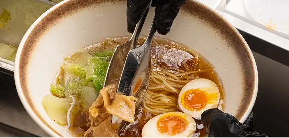 Hands wearing black gloves using tongs to add chicken to a bowl of ramen with half-boiled eggs, noodles, and leafy greens in broth.