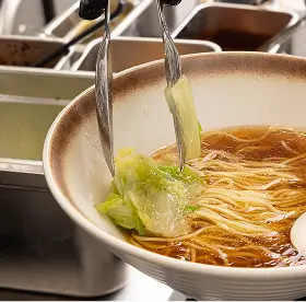 Chopsticks holding leafy greens over a bowl of noodle soup with broth and a white spoon.