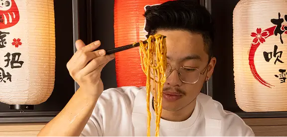 Man with glasses holding chopsticks lifting a portion of noodles in an Asian restaurant setting with decorative lanterns in the background.