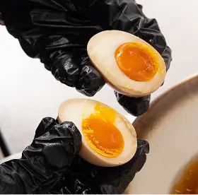 Hands wearing black gloves holding a ramen egg sliced in half showing its soft, bright orange yolk.
