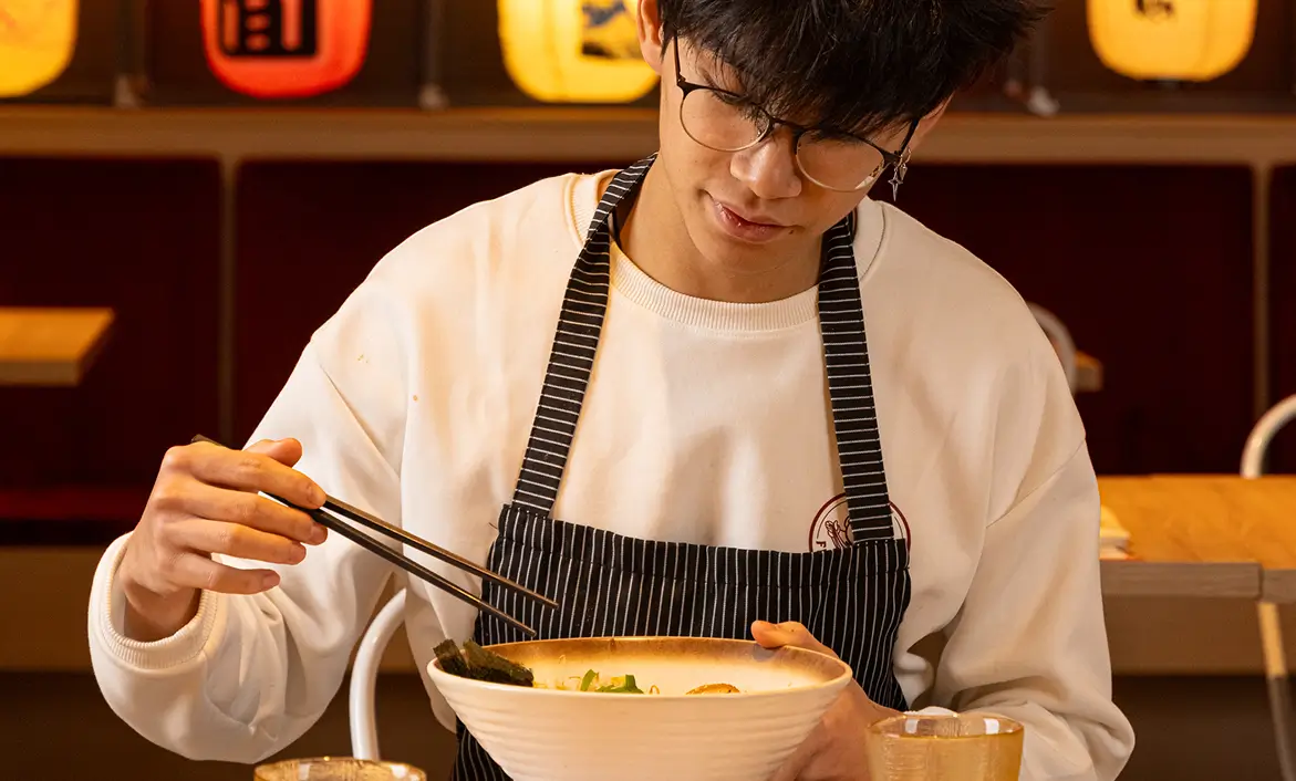 Person with glasses and striped apron eating noodles with chopsticks in a restaurant.