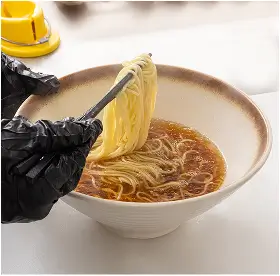 Person wearing black gloves lifting noodles with chopsticks from a bowl of broth.