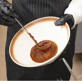 Person wearing black gloves and a pinstripe apron holding a white bowl with a thick swirl of dark reddish-brown sauce.