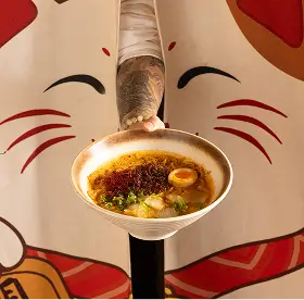 Close-up of a hand holding a bowl of ramen with egg, green onions, and chili oil, in front of a cartoon cat backdrop.