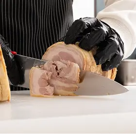 Person wearing black gloves slicing rolled roast meat with a large kitchen knife on a white surface.