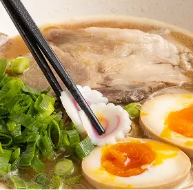 Chopsticks holding a pink and white flower-shaped fish cake over ramen with soft-boiled egg, green onions, and slices of pork.
