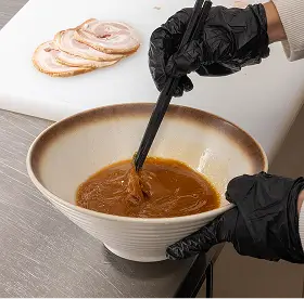 Person wearing black gloves stirring brown sauce in a bowl with chopsticks next to sliced cooked meat on a cutting board.