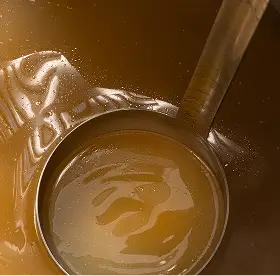 Close-up of a metal ladle holding smooth caramel or honey with a streaked swirl pattern inside a container.