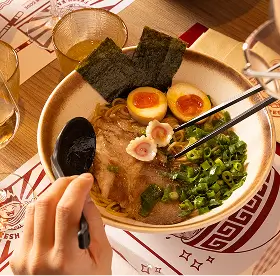 Bowl of ramen with sliced pork, soft-boiled eggs, seaweed, green onions, and noodles, held by chopsticks over a wooden table.