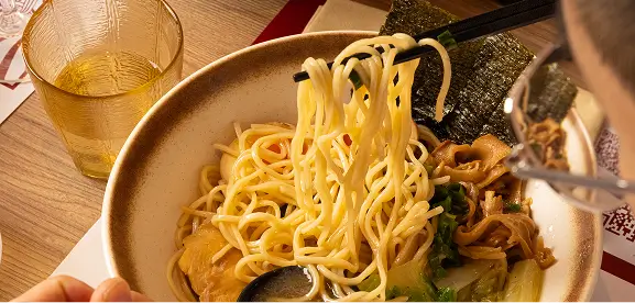 Close-up of a bowl of ramen noodles with chopsticks lifting noodles, accompanied by broth, vegetables, and a glass of tea nearby.