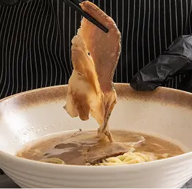 Thin slices of cooked meat being lifted with chopsticks above a bowl of noodle soup.