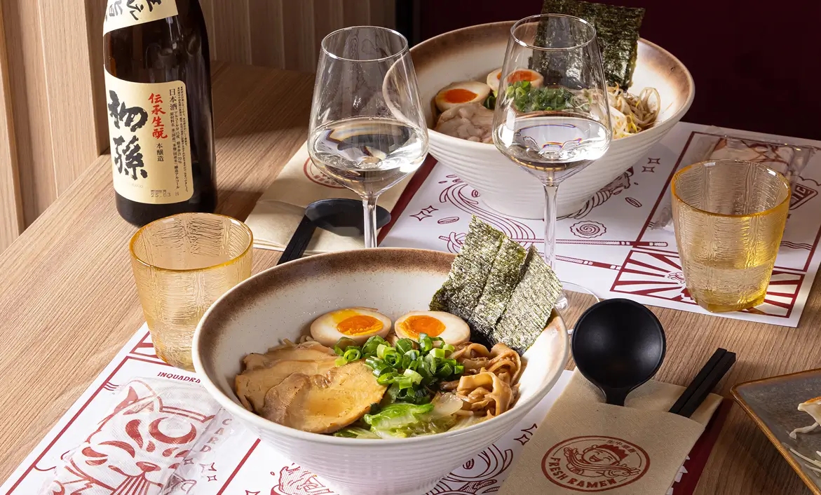 Two bowls of ramen with soft-boiled eggs, seaweed, green onions, and sliced pork on a wooden table with chopsticks, spoons, sake bottle, and glasses.