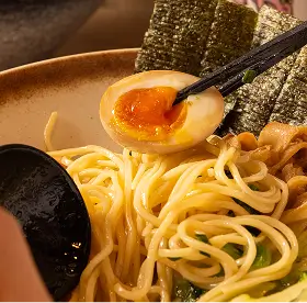 Close-up of ramen noodles with a halved soft-boiled egg held by chopsticks and seaweed sheets in the background.