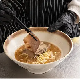 Person wearing black gloves and striped apron holding chopsticks over a bowl of ramen with broth and a slice of meat.