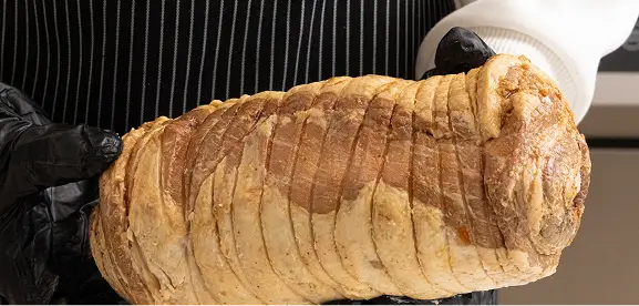 Person wearing black gloves holding a large piece of rolled, cooked meat with a browned outer crust.