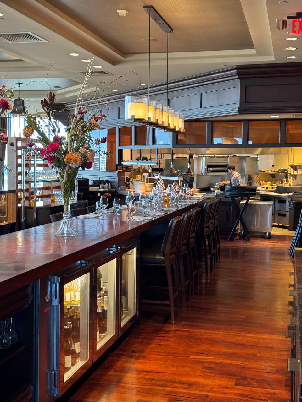 A chef working the pass behind an open kitchen counter lined with empty bar stools