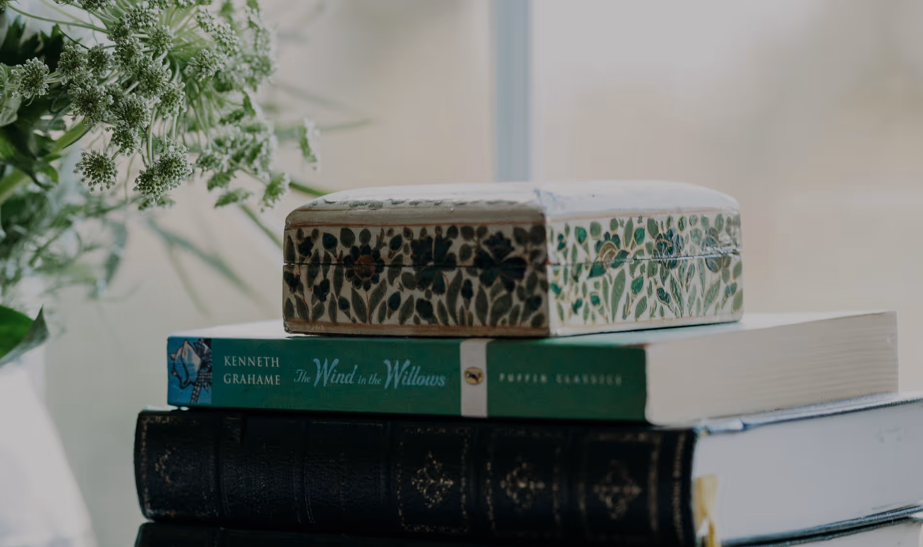 Stack of two books with a decorative floral box on top beside green leafy plant.