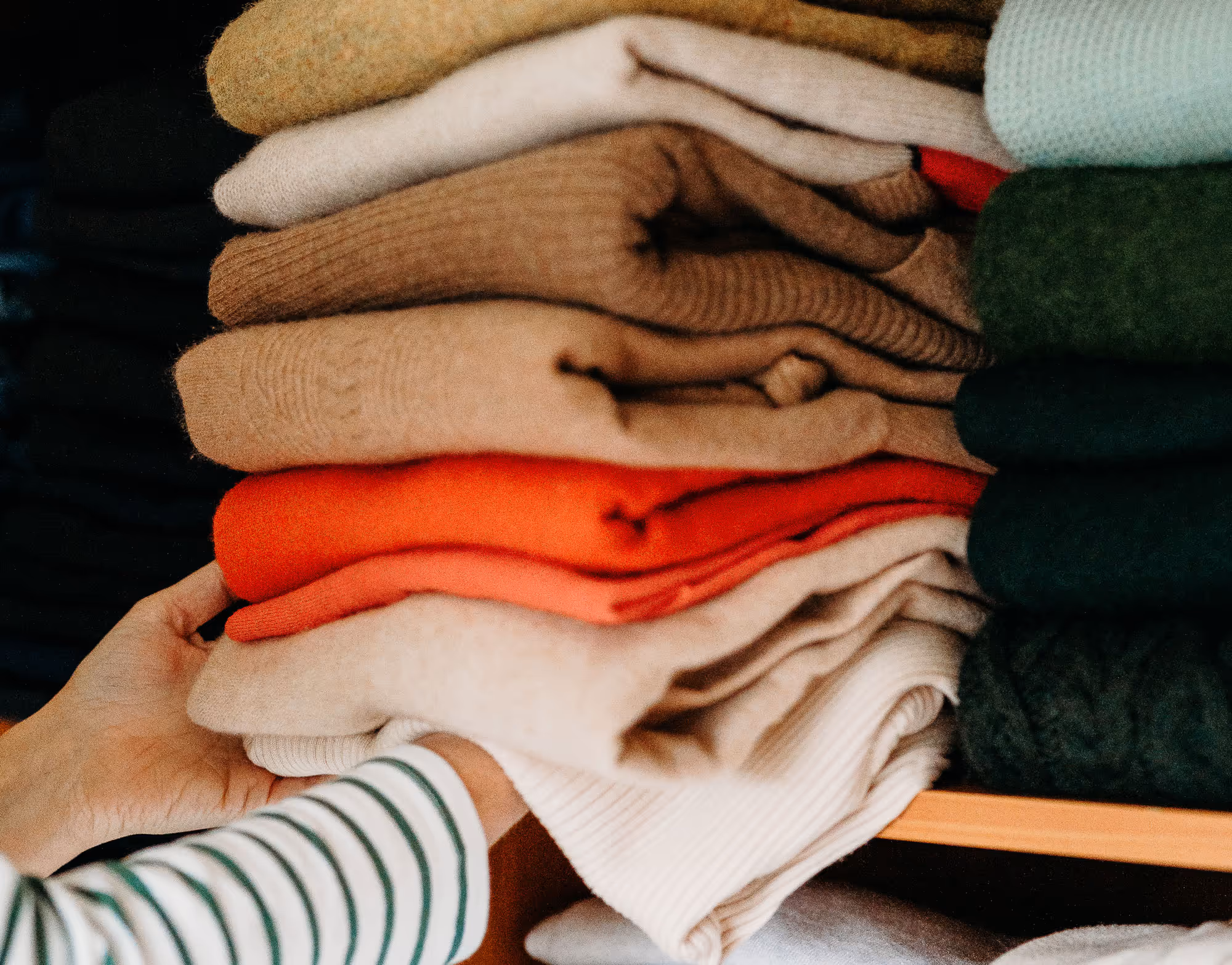 Hands organizing folded sweaters in various colors on a wooden shelf.