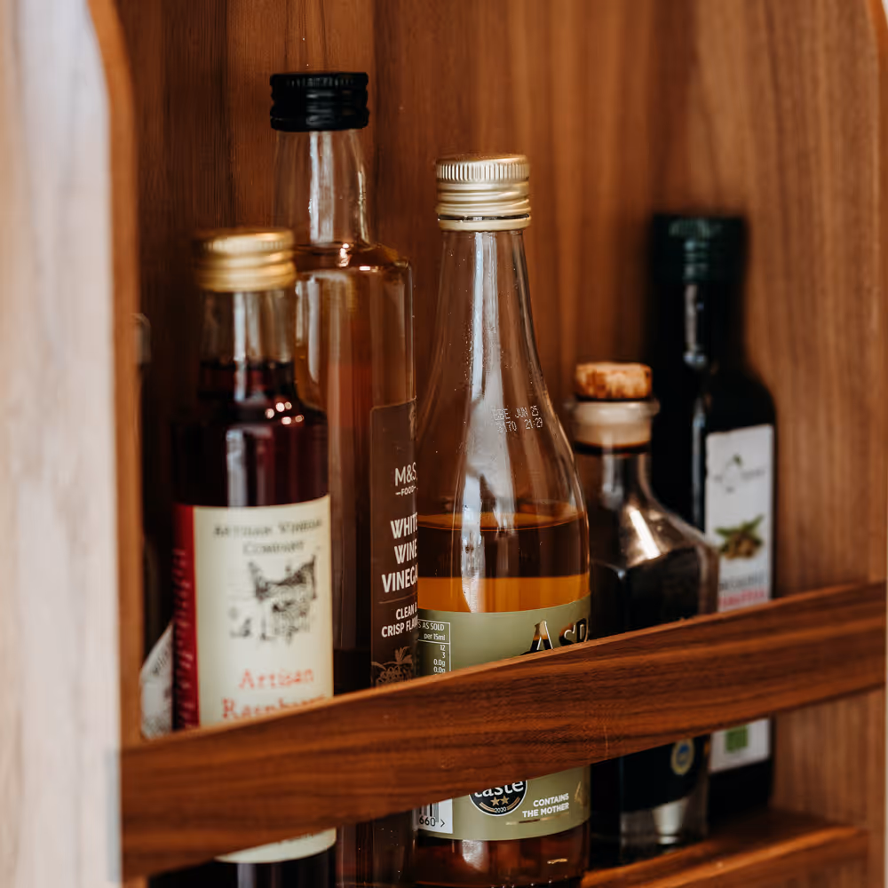 Wooden cupboard shelf holding five glass bottles of vinegar and oil with various labels and caps.