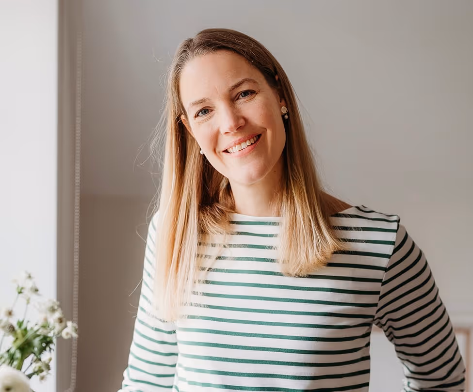 Smiling woman with straight blonde hair wearing a white and green striped shirt indoors near flowers.