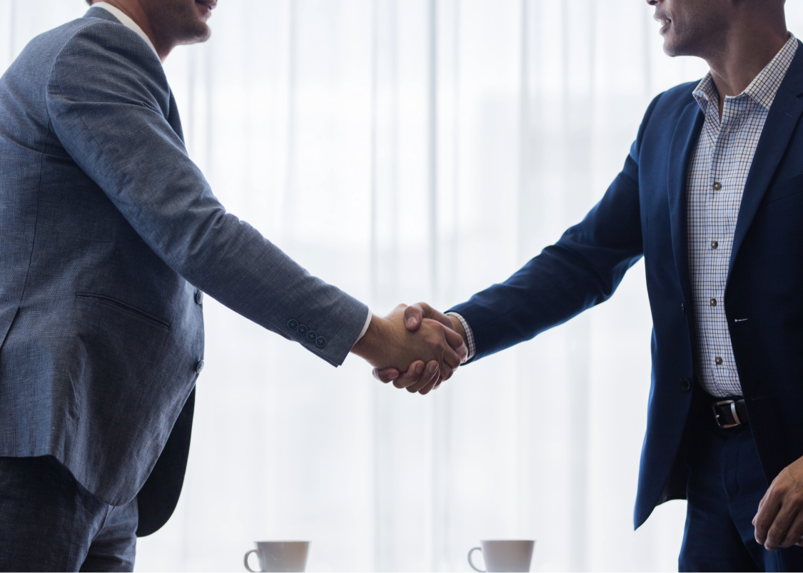 Two businessmen in suits shaking hands in front of bright sheer curtains with cups on table below.