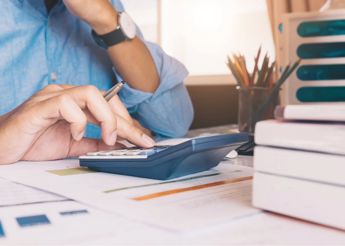 Person using calculator and holding pen on desk with papers, books, and pencil holder.