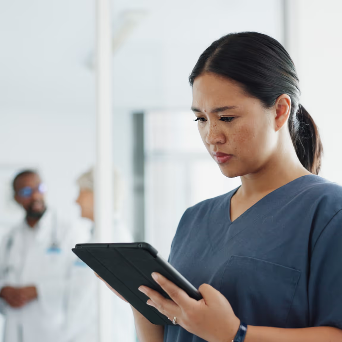 An Asian American female office manager in blue scrubs looking at a tablet, exuding a professional and approachable demeanor in a clinical setting.